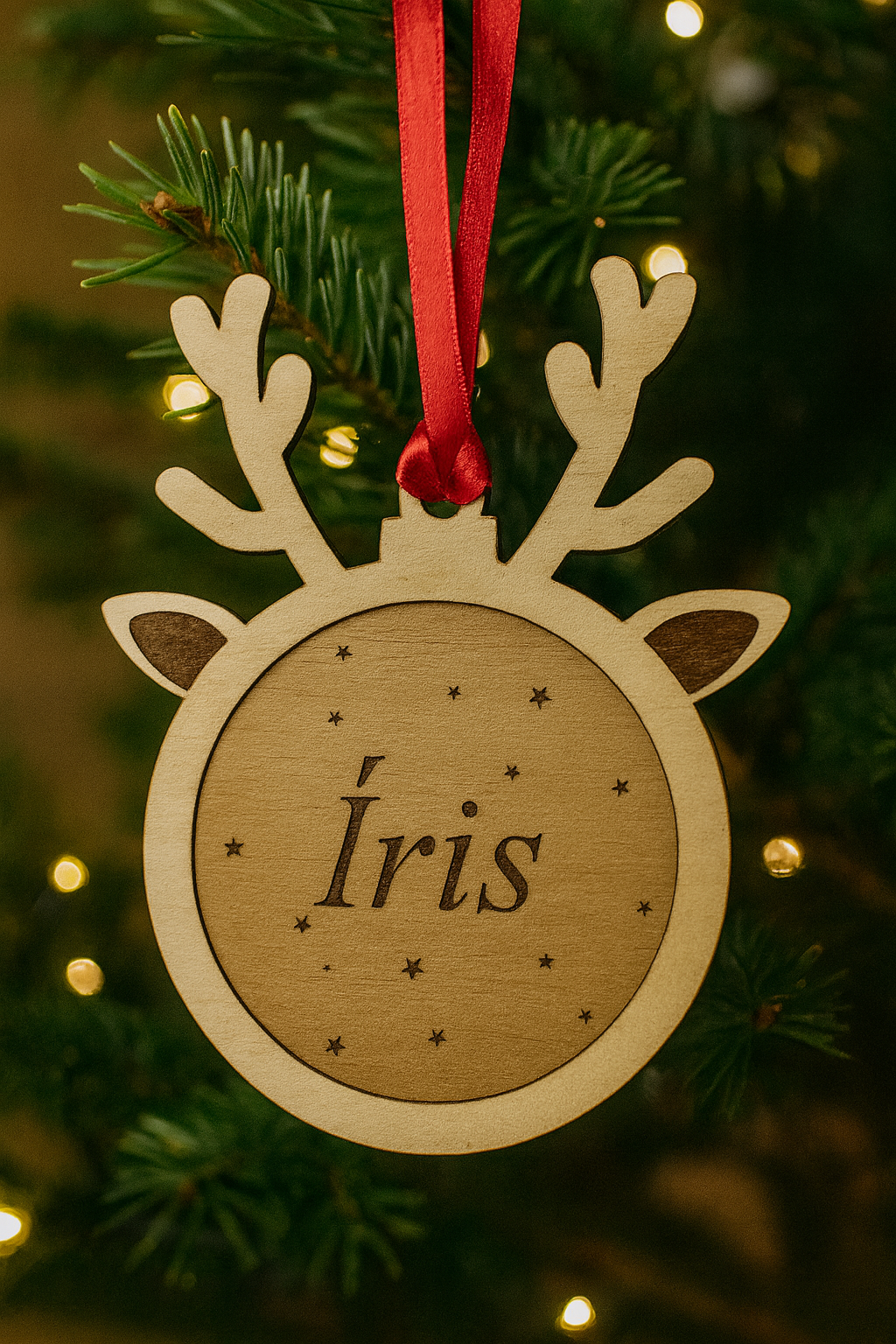 Bola de Natal personalizada em madeira de bétula, em formato de rena com fita de cetim vermelha, pendurada num pinheiro com luzes de Natal. Gravação a laser com o nome “Íris” e pequenas estrelas decorativas.