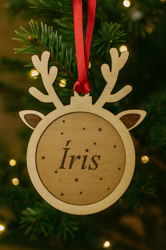 Bola de Natal personalizada em madeira de bétula, em formato de rena com fita de cetim vermelha, pendurada num pinheiro com luzes de Natal. Gravação a laser com o nome “Íris” e pequenas estrelas decorativas.