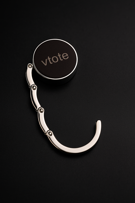 Cabide dobrável de metal com acabamento prateado e topo em couro PU castanho escuro, com gravação laser “Vtote”, segurando uma mala em mesa de restaurante moderna.